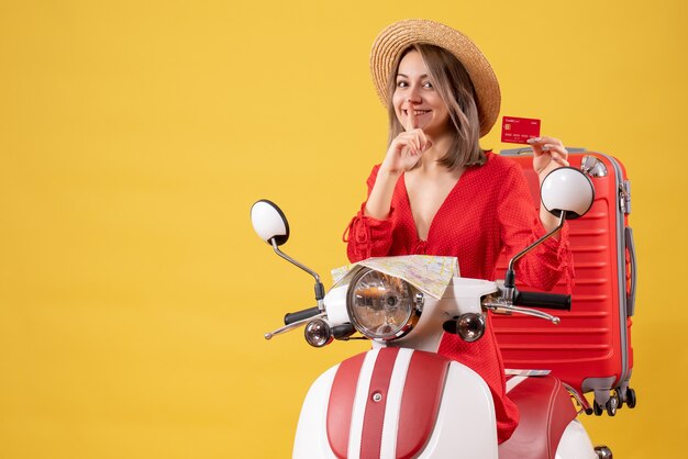 Vue de face jeune femme en robe rouge tenant une carte bancaire faisant signe chut sur cyclomoteur