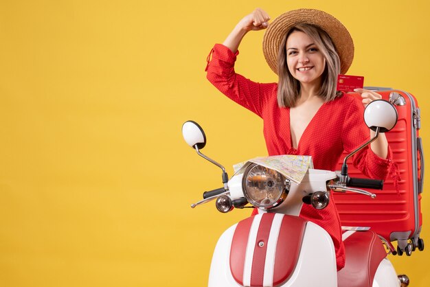 Vue de face jeune femme en robe rouge tenant une carte de réduction près d'un cyclomoteur