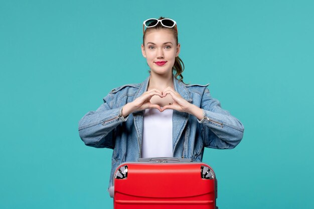 Vue de face jeune femme avec sac rouge se préparant pour les vacances et montrant un signe d'amour sur l'espace bleu