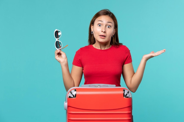 Vue de face jeune femme avec sac rouge tenant ses lunettes de soleil sur l'espace bleu clair