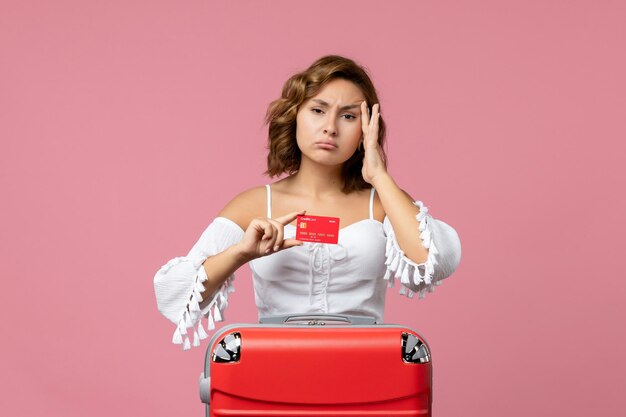 Vue de face d'une jeune femme avec un sac de vacances tenant une carte bancaire sur le mur rose