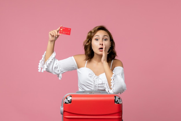 Vue de face d'une jeune femme avec un sac de vacances tenant une carte bancaire sur le mur rose