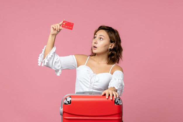 Vue de face d'une jeune femme avec un sac de vacances tenant une carte bancaire rouge sur le mur rose