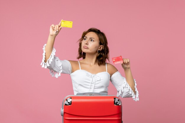 Vue de face d'une jeune femme avec un sac de vacances tenant des cartes bancaires sur un mur rose clair