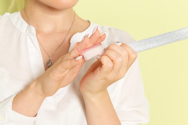 Vue de face jeune femme séduisante en chemise blanche fixant ses ongles sur une surface vert clair