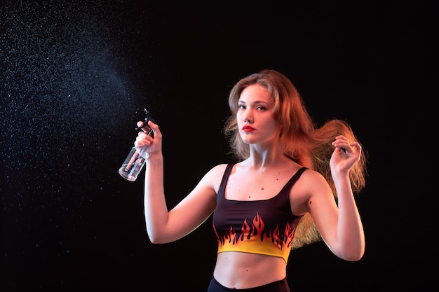 Une vue de face jeune femme séduisante en chemise de feu et pantalon noir à l'aide de spray de nettoyage sur le fond noir de nettoyage par pulvérisation