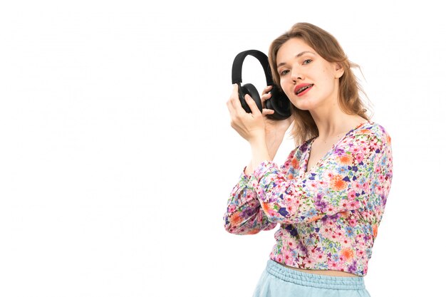 Une vue de face jeune femme séduisante en chemise à fleurs colorées et jupe bleue tenant des écouteurs noirs sur le blanc