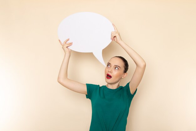 Photo gratuite vue de face jeune femme séduisante en chemise vert foncé et jean bleu tenant une pancarte blanche sur beige