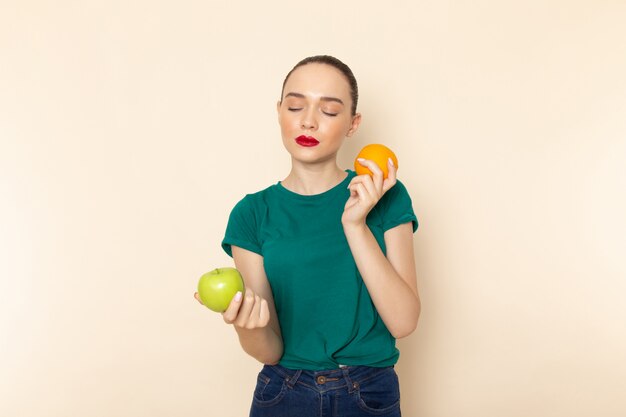 Vue de face jeune femme séduisante en chemise vert foncé tenant orange et pomme avec les yeux fermés sur beige