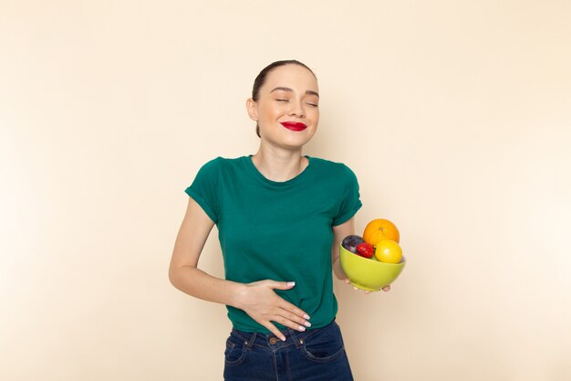 Vue de face jeune femme séduisante en chemise vert foncé tenant la plaque avec des fruits avec sourire