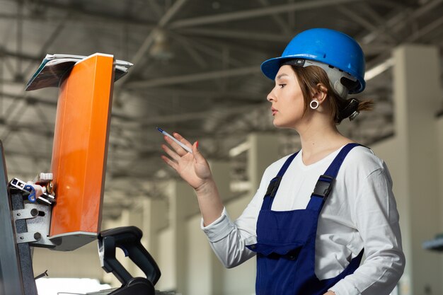 Une vue de face jeune femme séduisante en costume de construction bleu et casque contrôlant les machines dans le hangar pendant la construction de l'architecture des bâtiments de jour