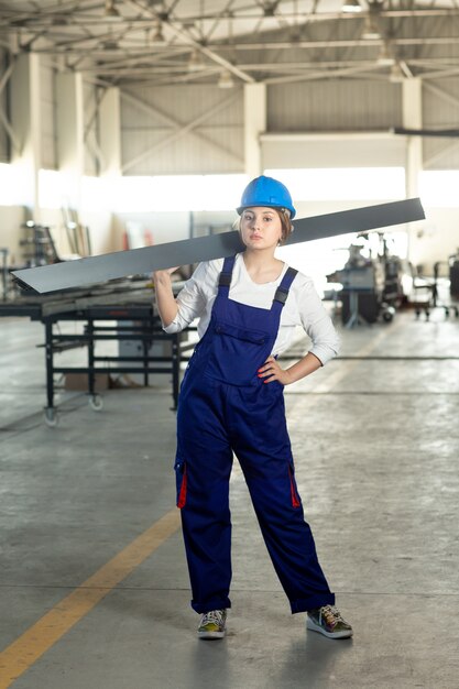 Une vue de face jeune femme séduisante en costume de construction bleu et casque de travail tenant des détails métalliques lourds pendant la construction de l'architecture des bâtiments pendant la journée