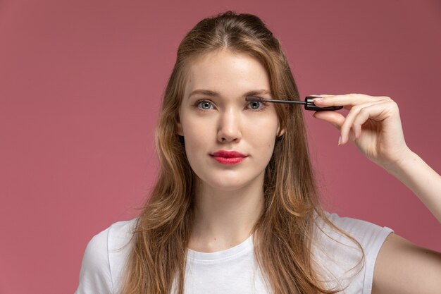 Vue de face jeune femme séduisante faisant son maquillage avec du mascara sur mur rose foncé couleur modèle femme jeune fille