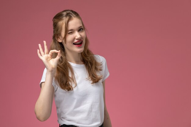 Vue de face jeune femme séduisante posant avec sourire et clin d'oeil sur le modèle de mur rose couleur jeune femme