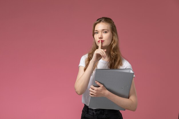 Vue de face jeune femme séduisante en t-shirt blanc demandant d'être silencieux tenant document gris sur mur rose modèle couleur femme jeune fille