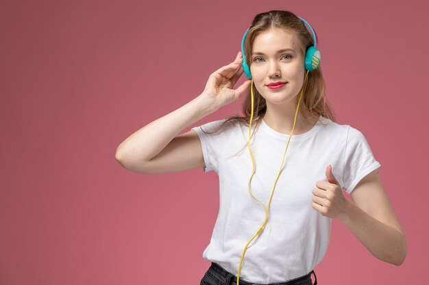 Vue de face jeune femme séduisante en t-shirt blanc, écouter de la musique sur le modèle de bureau rose foncé couleur jeune fille