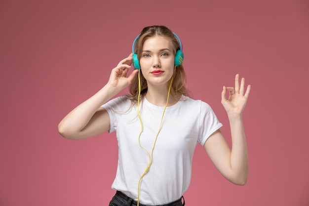 Vue de face jeune femme séduisante en t-shirt blanc, écouter de la musique avec ses écouteurs sur le modèle de bureau rose couleur femme jeune fille