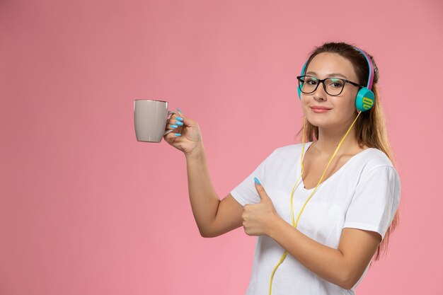 Vue de face jeune femme séduisante en t-shirt blanc, écouter de la musique tenant une tasse en souriant sur le fond rose
