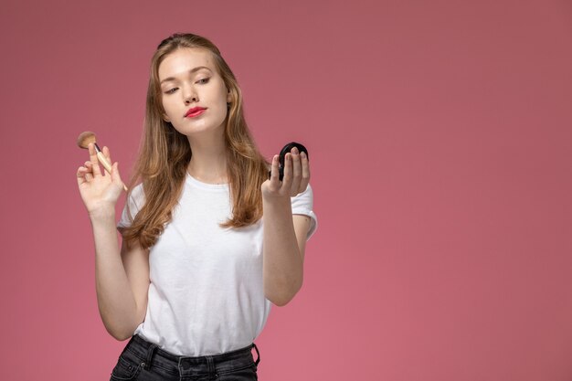 Vue de face jeune femme séduisante en t-shirt blanc faisant du maquillage sur le mur rose modèle couleur jeune femme