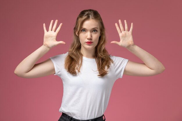 Vue de face jeune femme séduisante en t-shirt blanc avec les mains levées posant sur le mur rose modèle couleur jeune femme