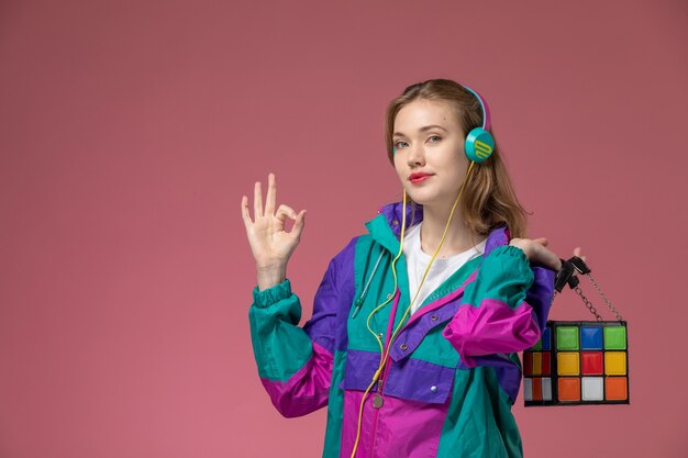 Vue de face jeune femme séduisante en t-shirt blanc manteau coloré écouter de la musique via des écouteurs avec sourire sur le mur rose modèle femme pose photo couleur