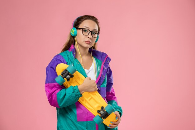 Vue de face jeune femme séduisante en t-shirt blanc manteau coloré posant avec planche à roulettes sur fond rose