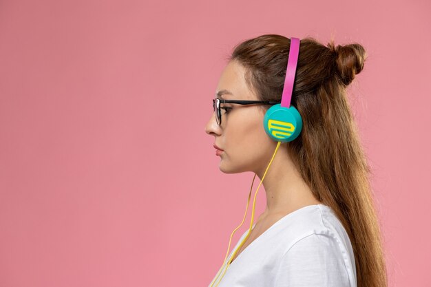 Vue de face jeune femme séduisante en t-shirt blanc posant et écoutant de la musique via un casque sur le fond rose