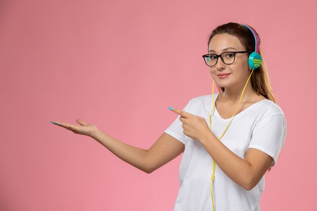 Vue de face jeune femme séduisante en t-shirt blanc posant et écoutant de la musique via des écouteurs souriant sur le fond rose