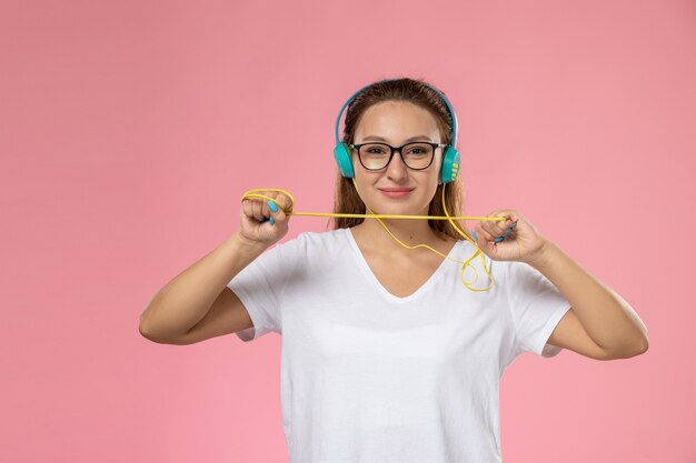 Vue de face jeune femme séduisante en t-shirt blanc posant et écoutant de la musique via des écouteurs avec un sourire léger sur le fond rose