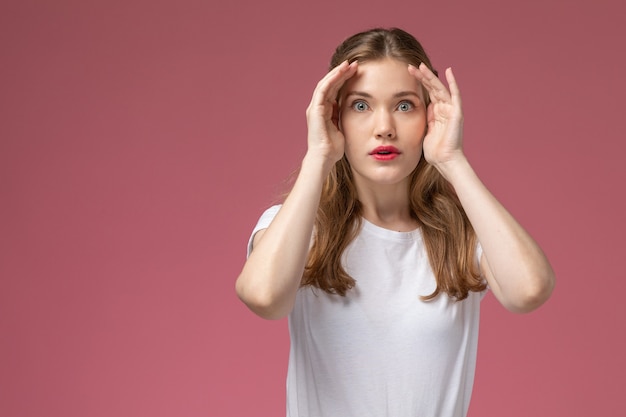 Vue de face jeune femme séduisante en t-shirt blanc posant avec une expression surprise sur le modèle de mur rose femme pose photo couleur