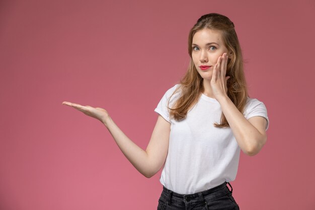 Vue de face jeune femme séduisante en t-shirt blanc posant avec une expression timide sur le modèle de mur rose foncé couleur femme jeune fille