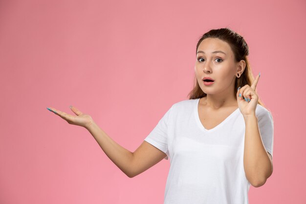 Vue de face jeune femme séduisante en t-shirt blanc posant avec la main levée et expression surprise sur fond rose