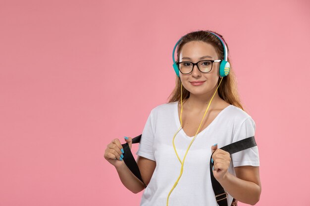 Vue de face jeune femme séduisante en t-shirt blanc posant et smi écouter de la musique sur fond rose