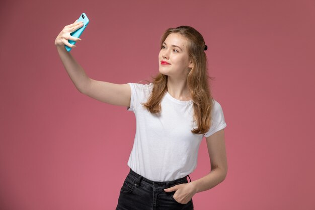 Vue de face jeune femme séduisante en t-shirt blanc prenant un selfie sur mur rose modèle couleur femme fille