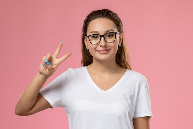 Vue de face jeune femme séduisante en t-shirt blanc smi et montrant le signe de la victoire sur le fond rose