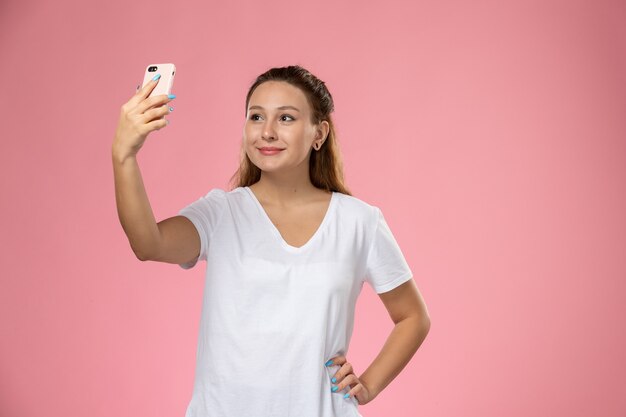 Vue de face jeune femme séduisante en t-shirt blanc avec sourire en prenant un selfie sur le fond rose