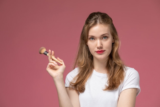 Vue de face jeune femme séduisante en t-shirt blanc tenant un pinceau de maquillage sur le mur rose modèle couleur jeune femme