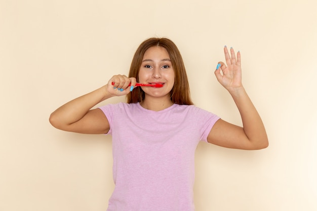 Vue de face jeune femme séduisante en t-shirt rose et blue-jeans nettoyant ses dents avec sourire