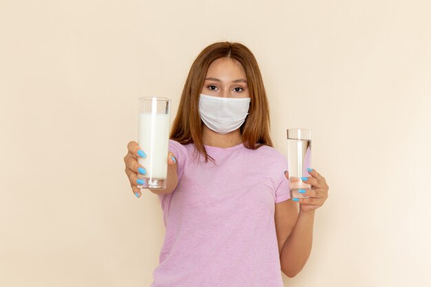 Vue de face jeune femme séduisante en t-shirt rose et blue-jeans tenant des verres d'eau et de lait avec masque