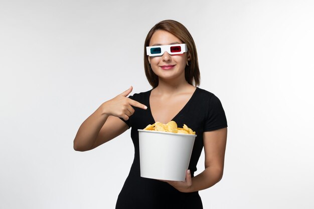 Vue de face jeune femme séduisante tenant le panier avec des cips de pommes de terre dans des lunettes de soleil sur une surface blanche