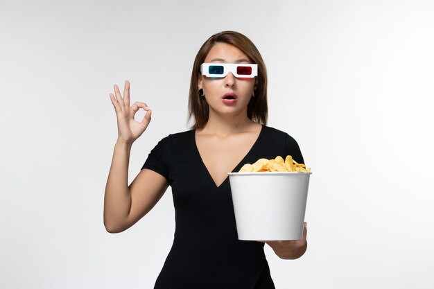 Vue de face jeune femme séduisante tenant panier avec pommes de terre cips dans des lunettes de soleil sur une surface blanche