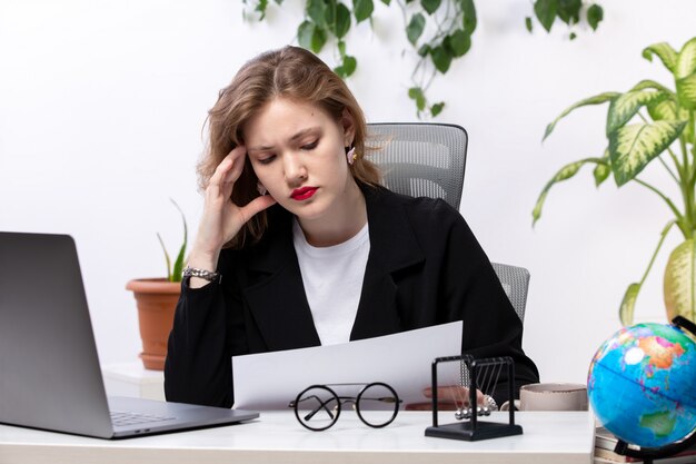 Une vue de face jeune femme séduisante en veste noire et chemise blanche en face de la table de travail avec des documents d'ordinateur portable technologies de travail