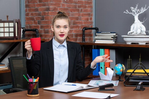 Vue de face d'une jeune femme souriante assise à une table et tenant une tasse rouge au bureau