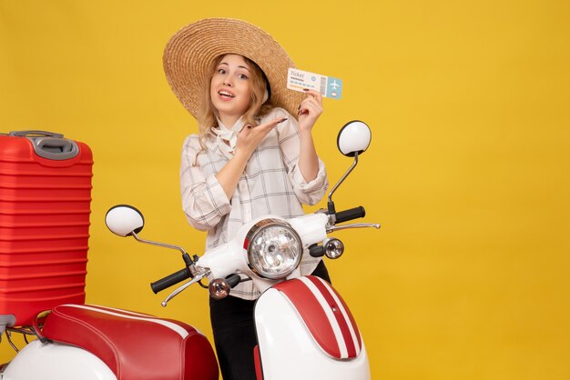 Vue de face de la jeune femme souriante portant un chapeau rassemblant ses bagages assis sur la moto et montrant le billet