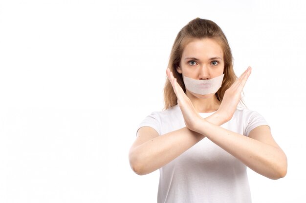 Une vue de face jeune femme en t-shirt blanc avec un bandage blanc autour de sa bouche montrant le signe d'interdiction sur le blanc