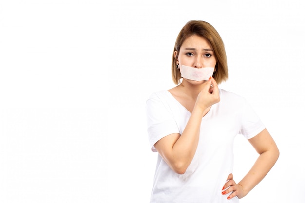 Une vue de face jeune femme en t-shirt blanc avec un bandage blanc autour de sa bouche en touchant son bandage sur le blanc