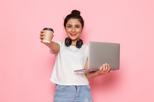 Une vue de face jeune femme en t-shirt blanc et blue-jeans tenant un ordinateur portable et une tasse de café