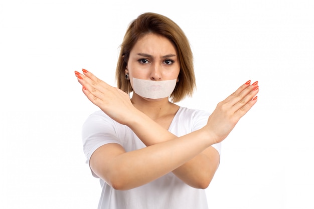 Une vue de face jeune femme en t-shirt blanc et jean noir portant un bandage blanc autour de sa bouche montrant le signe d'interdiction sur le blanc