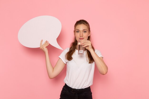 Vue de face de la jeune femme en t-shirt blanc tenant une pancarte blanche et un verre d'eau sur le mur rose