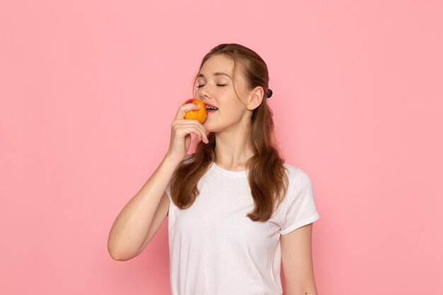 Vue de face de la jeune femme en t-shirt blanc tenant la pêche fraîche le manger sur un mur rose clair